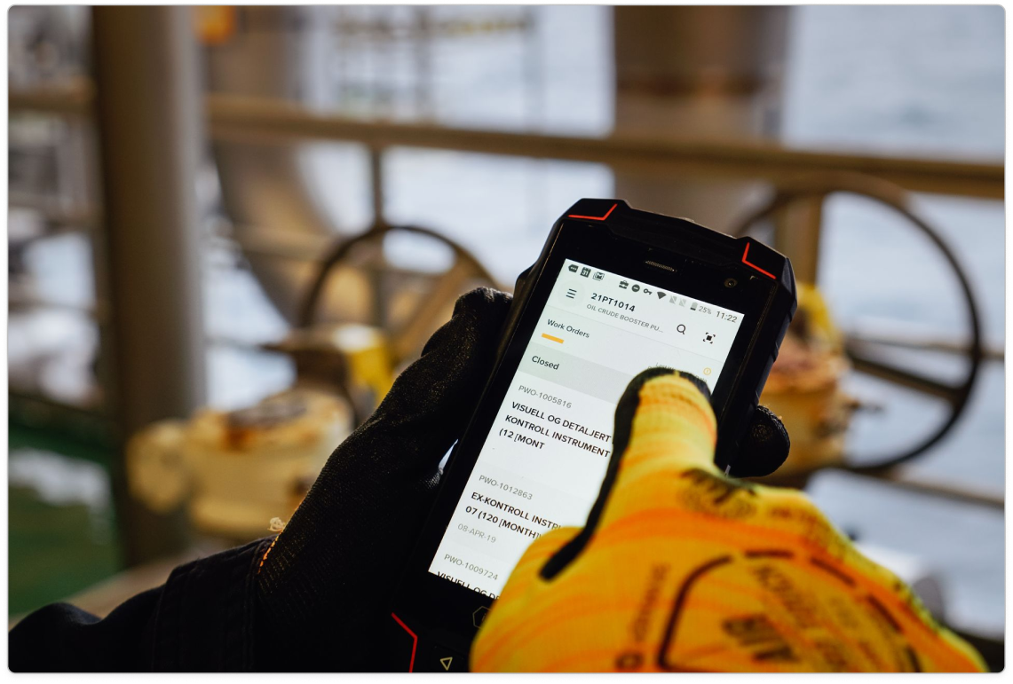 Gloved hand holding a rugged phone showing a work orders list on an offshore platform
